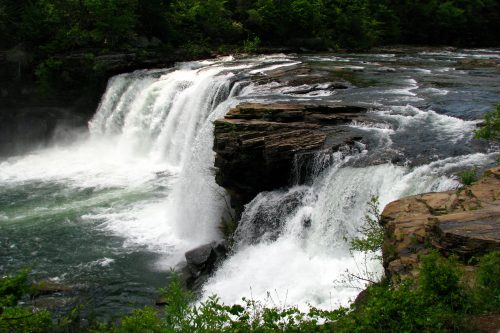 A cascading waterfall flows over rocky cliffs, surrounded by lush green trees and a serene river.