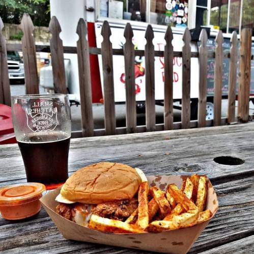 A burger with fries and dipping sauce on a wooden table, with a glass of beer and a food truck in the background.