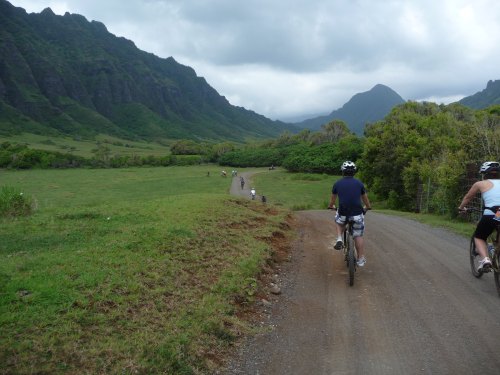 A group of cyclists rides along a dirt path surrounded by lush green hills and mountains under a cloudy sky.