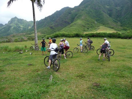A group of mountain bikers gathers in a grassy area with lush green mountains in the background.