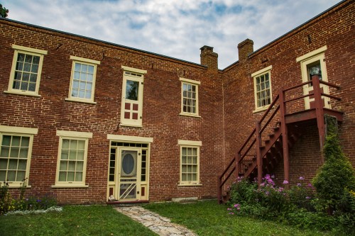 A brick building with a wooden staircase, surrounded by greenery and flowers, under a cloudy sky.