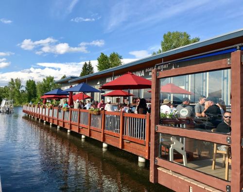 A waterfront restaurant with outdoor seating, umbrellas, and diners enjoying the view along a calm waterway.