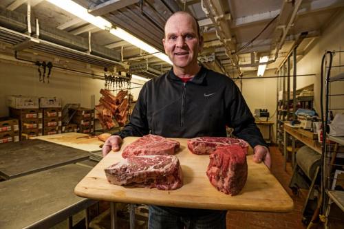 A man holds a wooden board with large cuts of meat in a butcher shop, surrounded by meat storage and equipment.