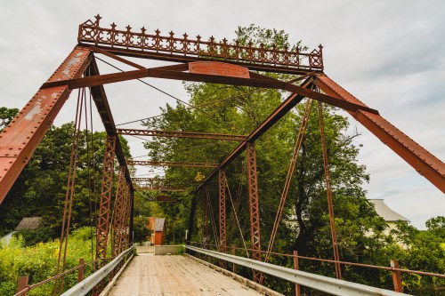 Rusty metal bridge with decorative arches, surrounded by greenery and a cloudy sky.