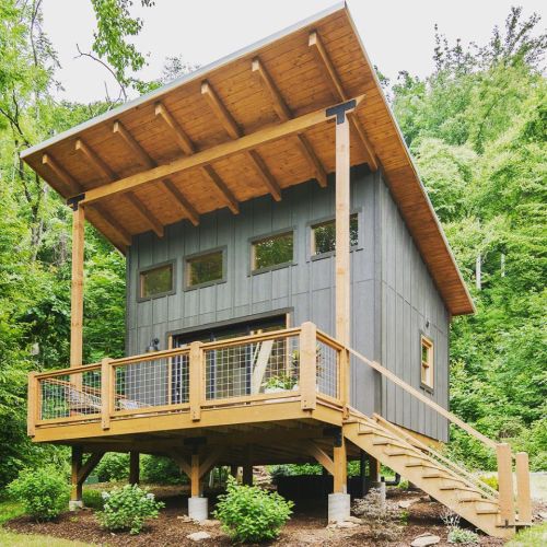 A modern cabin with a wooden deck, elevated on stilts, surrounded by lush greenery and trees.