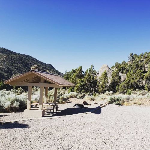 A picnic shelter in a gravel area surrounded by trees and mountains under a clear blue sky.