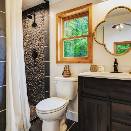 Modern bathroom featuring a pebble shower, wooden accents, and a stylish mirror, with natural light from a window.