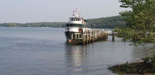A boat docked at a wooden pier on a calm river, surrounded by green trees and a clear sky.