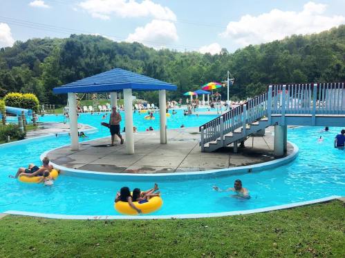 A sunny pool scene with people swimming and relaxing on inflatable tubes, surrounded by greenery and colorful umbrellas.