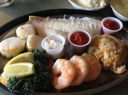 A seafood platter featuring grilled fish, scallops, shrimp, lemon wedges, and dipping sauces on a metal tray.