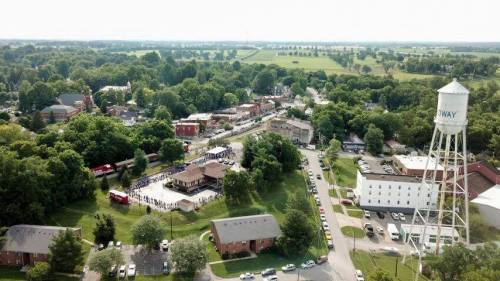 Aerial view of a small town with a water tower, green fields, and a gathering of people near a building.