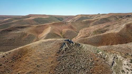 Aerial view of rolling, dry hills with sparse vegetation under a clear blue sky.