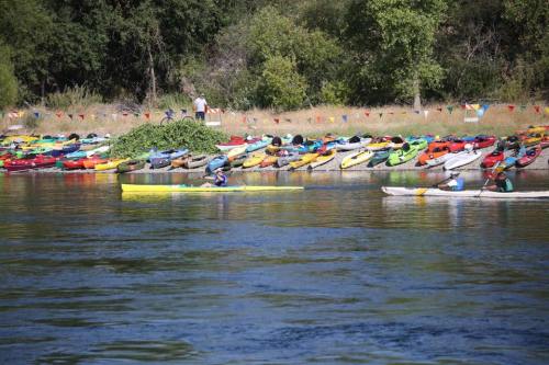 A serene river scene with kayakers paddling past a colorful array of parked kayaks along the shore.