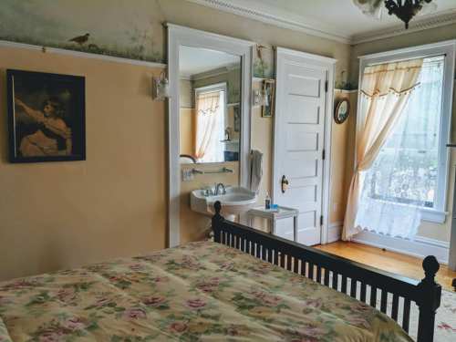 Cozy bedroom with floral bedding, a mirror, and a window draped with sheer curtains, featuring vintage decor.
