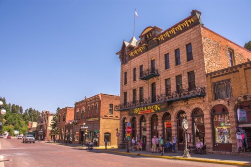 Historic buildings line a sunny street in a small town, featuring shops and a prominent hotel with a flag.