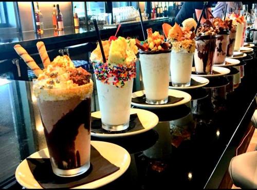 A row of colorful milkshakes topped with whipped cream, sprinkles, and various treats on a bar counter.