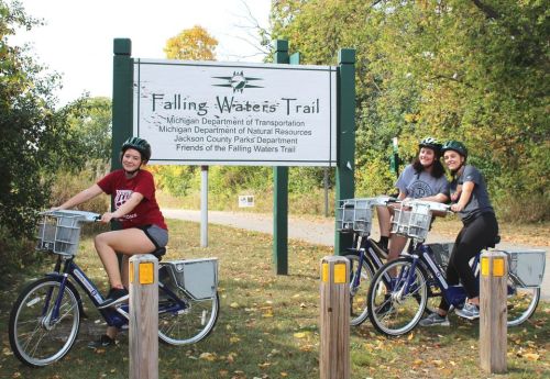 Three women in helmets pose on bicycles in front of the Falling Waters Trail sign, surrounded by greenery.