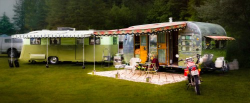 A vintage camper and a green food truck parked on grass, with outdoor seating and a motorcycle nearby.