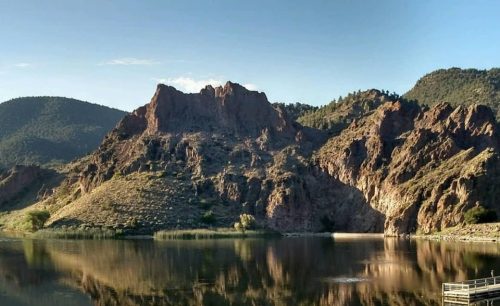 A serene lake reflects rugged mountains and greenery under a clear sky. A wooden dock is visible in the foreground.