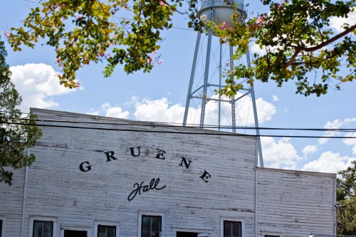 Historic Gruene Hall building with a water tower in the background, framed by tree branches and a blue sky.
