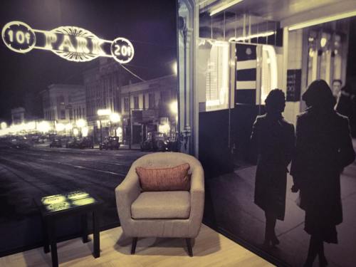 A cozy corner with a gray armchair and a table, featuring vintage black-and-white wall art of a city street and figures.