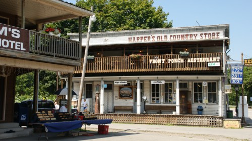 A rustic country store with a porch, featuring a restaurant sign and an ATM, surrounded by trees and a small town atmosphere.