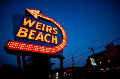 Neon sign for Weirs Beach illuminated at dusk, with a red arrow and bright lights against a darkening sky.