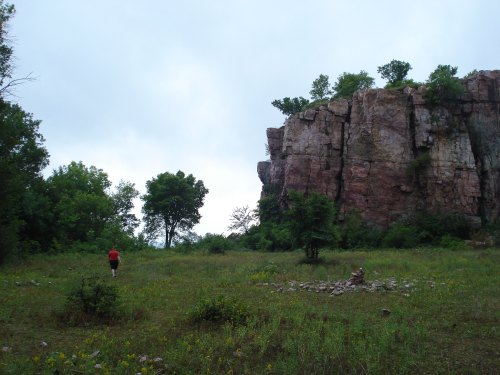A person walks through a grassy area near a large rock formation and trees under a cloudy sky.