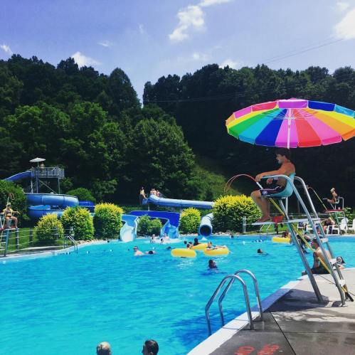 A sunny pool scene with people swimming, a lifeguard on duty, and colorful slides in the background.