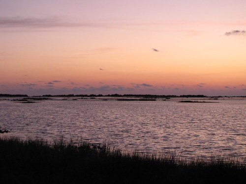 A serene sunset over a calm body of water, with silhouettes of distant land and soft clouds in the sky.