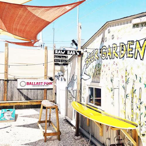 A sunny outdoor space featuring a beer garden, raw bar sign, and a surfboard against a colorful mural.