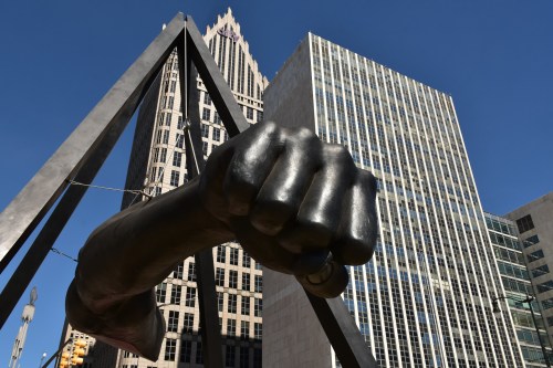 A large bronze fist sculpture stands against a backdrop of modern buildings and a clear blue sky.