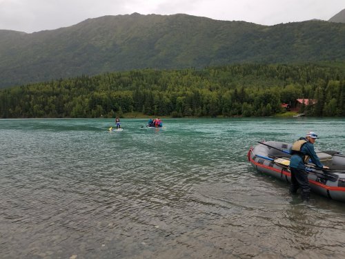 Two people paddle in a kayak on a calm river, while another person prepares a raft on the shore, surrounded by mountains.