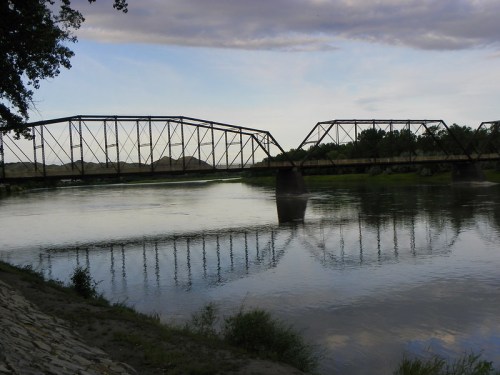 A metal bridge spans a calm river, reflecting in the water under a cloudy sky. Lush greenery lines the riverbank.