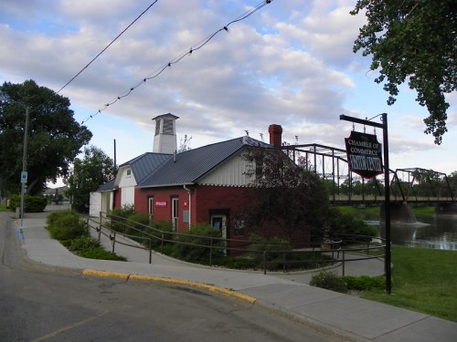 Historic building with a red brick facade, a metal roof, and a sign for the Chamber of Commerce by a river.