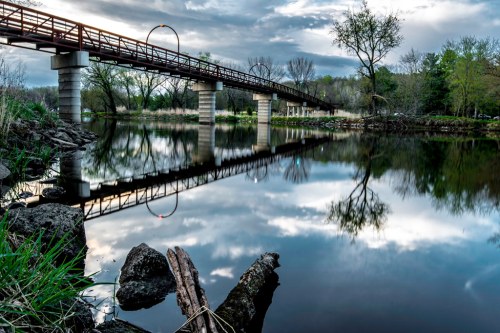 A wooden bridge spans a calm river, reflecting clouds and trees, with rocks and grass in the foreground.