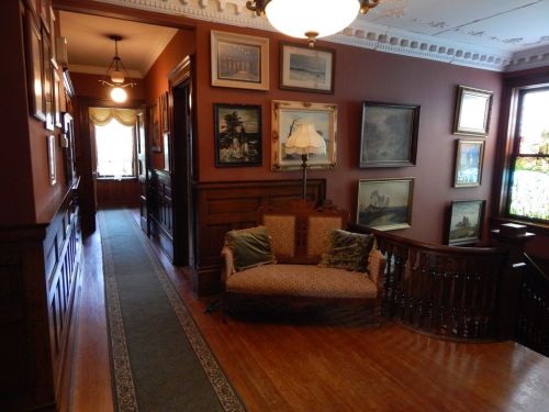 A hallway with wooden walls, framed artwork, a vintage lamp, and a patterned sofa, leading to a bright room.