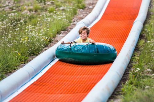 A young boy smiles as he rides a green inner tube down a bright orange slide surrounded by grass and flowers.
