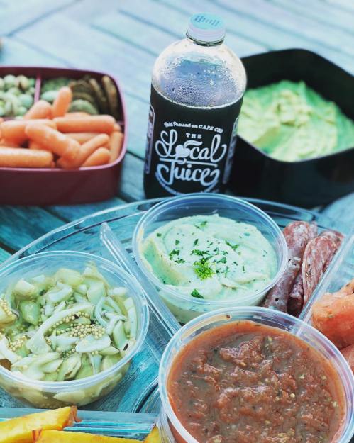 A colorful spread of fresh vegetables, dips, and a drink labeled "The Local Juice" on a wooden table.