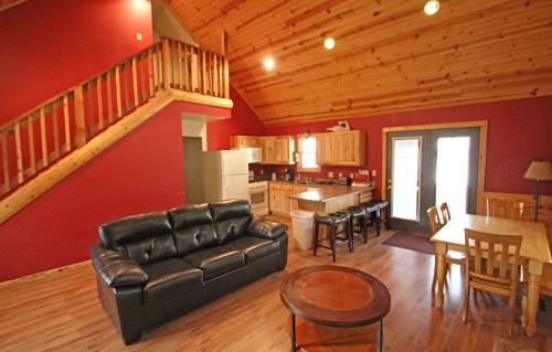 Cozy cabin interior with wooden accents, a black sofa, dining table, and a staircase leading to an upper level.
