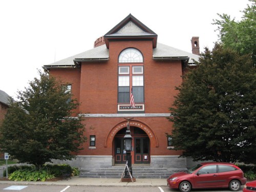 Historic brick city hall building with large windows, an American flag, and trees in front. A red car is parked nearby.