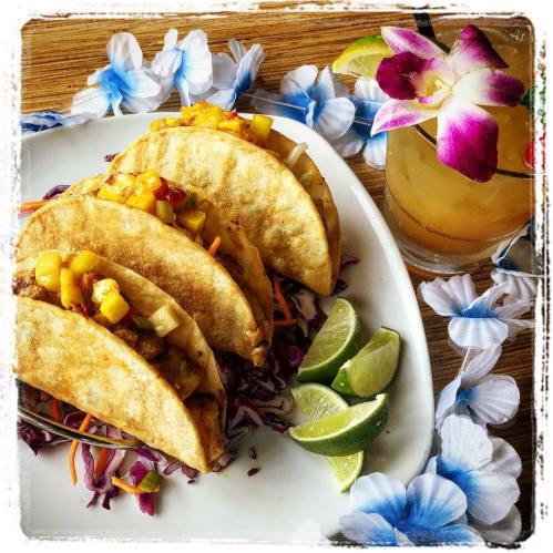 Three tacos filled with toppings, served on a plate with lime wedges and a tropical drink, surrounded by flower decorations.