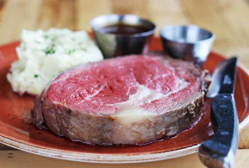 A plate with a juicy slice of prime rib, creamy mashed potatoes, and two small cups of sauce, accompanied by a knife.