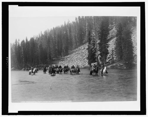 A historical black-and-white photo of a group crossing a river on horseback, surrounded by trees and rocky terrain.