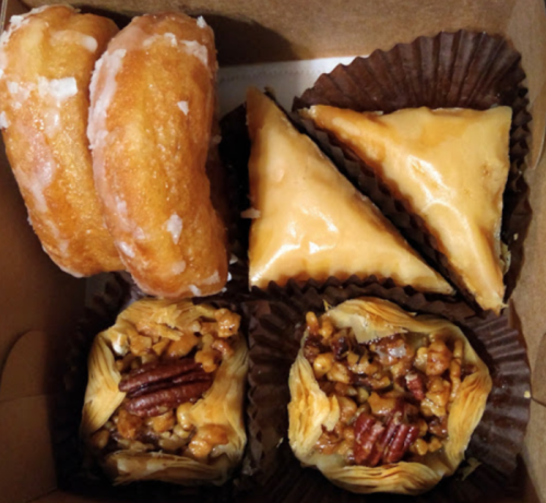 A box of assorted pastries, including glazed donuts, triangular baklava, and nut-filled phyllo cups.
