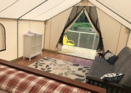 Cozy tent interior with a futon, rug, and a view of a deck by the water. Bright yellow chair outside.
