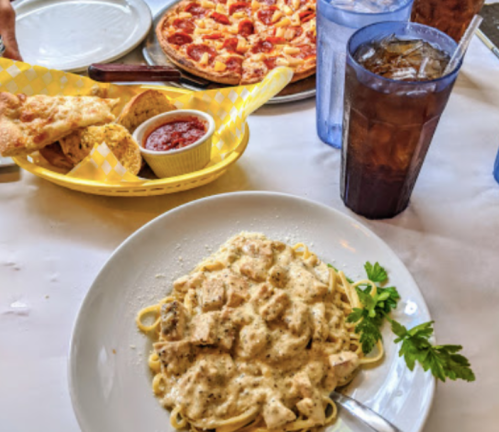 A plate of creamy chicken pasta with parsley, a basket of breadsticks, and a pizza in the background, with drinks.