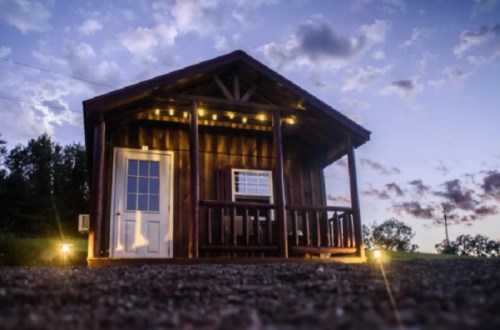 A small wooden cabin with a porch, illuminated by lights, set against a twilight sky with scattered clouds.