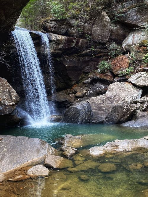 A serene waterfall cascades over rocks into a clear pool, surrounded by lush greenery and rugged stone formations.
