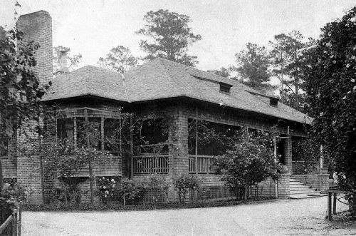 Historic brick house with a large porch, surrounded by trees and gardens, in a black-and-white photograph.